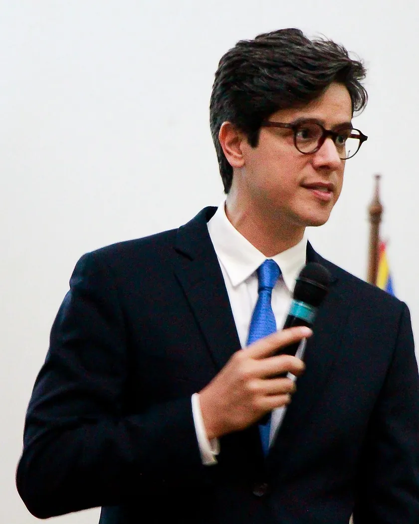 Leonardo Fogaça, de terno, falando ao microfone em um evento, com bandeiras ao fundo